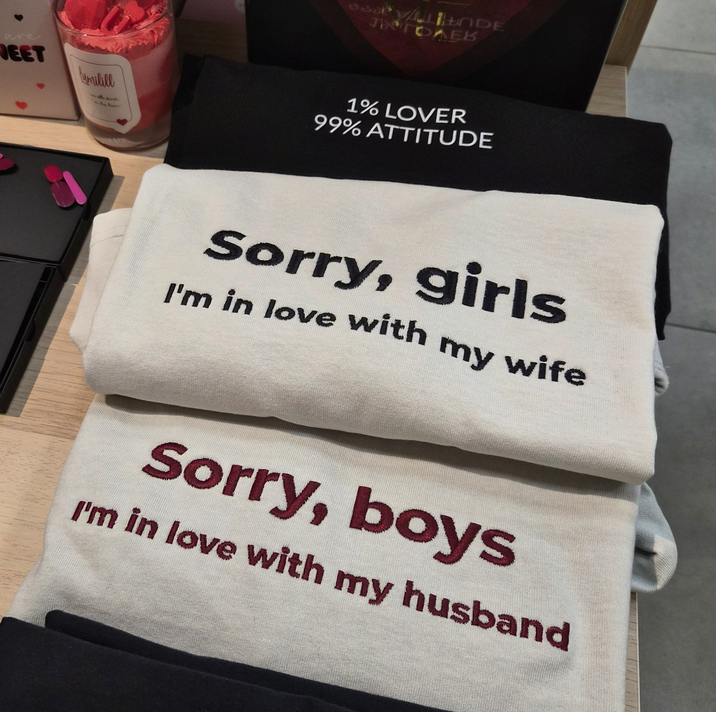 A stack of folded graphic t-shirts displayed on a wooden table. The top folded shirt is black with white text reading '1% LOVER 99% ATTITUDE'.