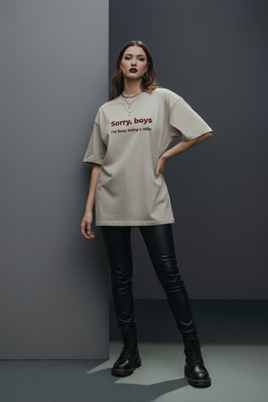 Person wearing a beige oversized t-shirt with text, black pants, and black boots against a gray background
